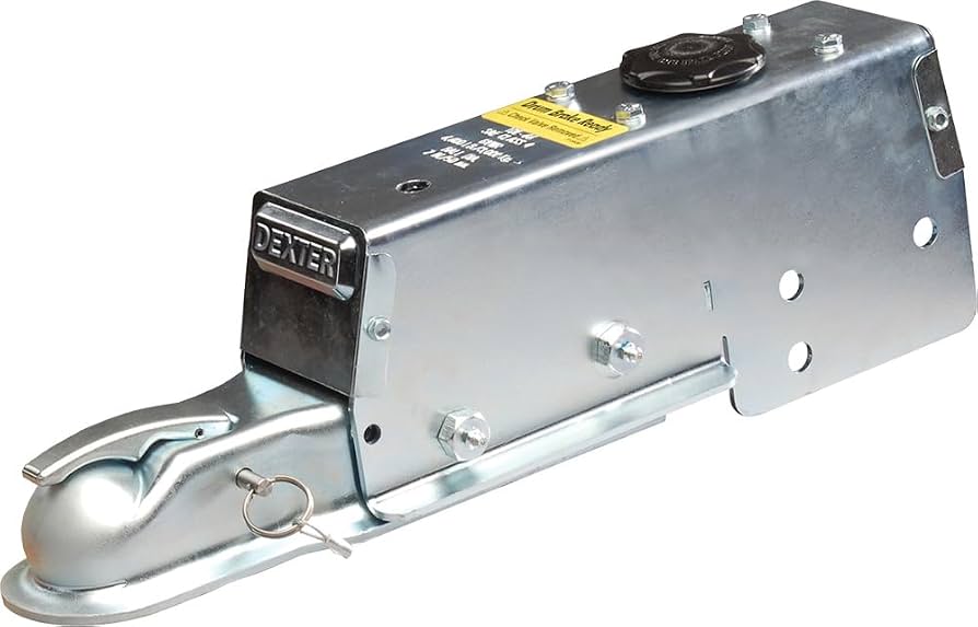 A close-up photograph of a hydraulic surge brake actuator mounted on a trailer tongue, showing the coupler housing, breakaway cable, and hydraulic brake line connection in daylight