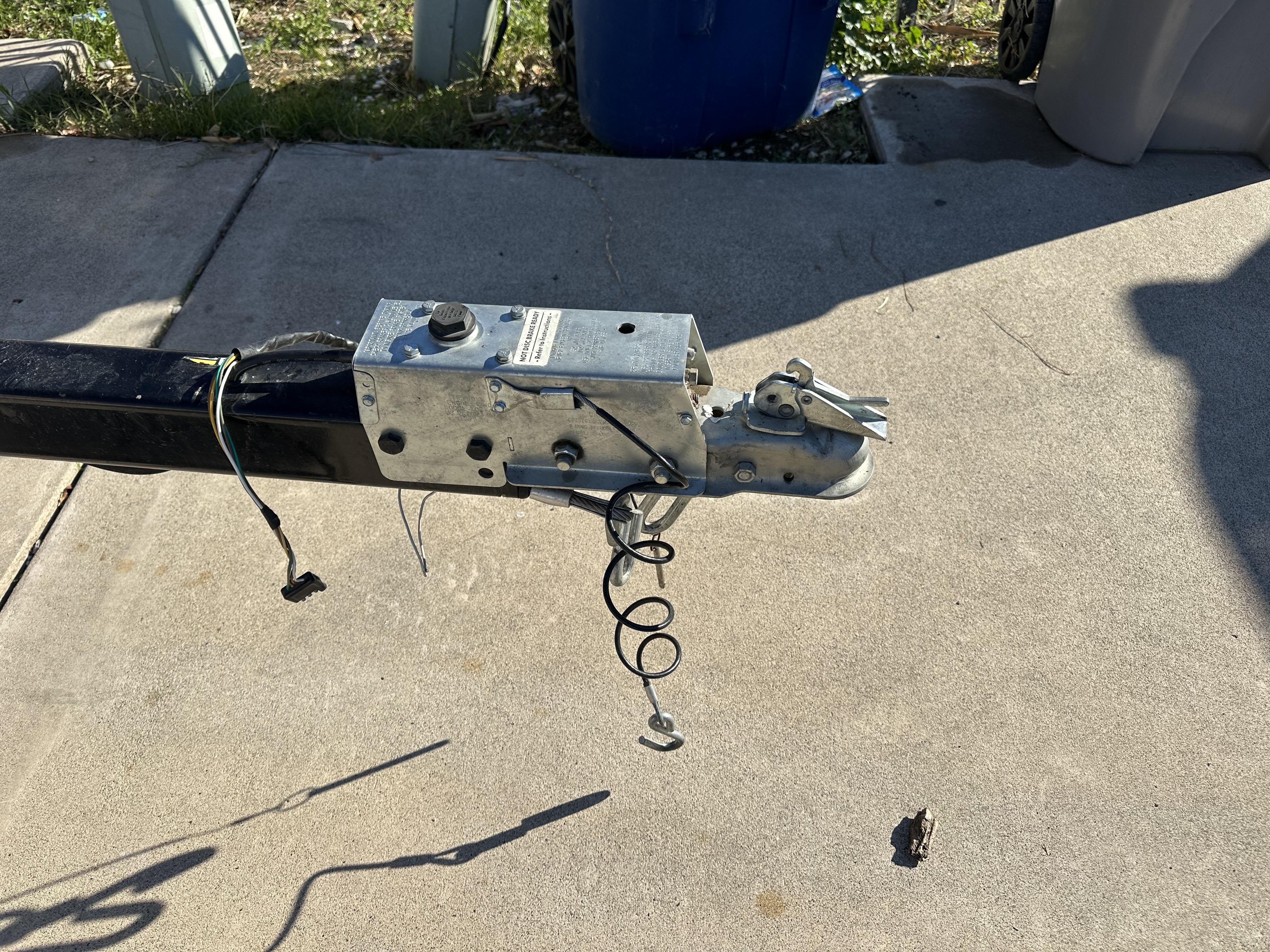 A photograph of a hydraulic surge brake coupler on a utility trailer tongue parked on a concrete driveway, with the hitch ball and safety chains visible