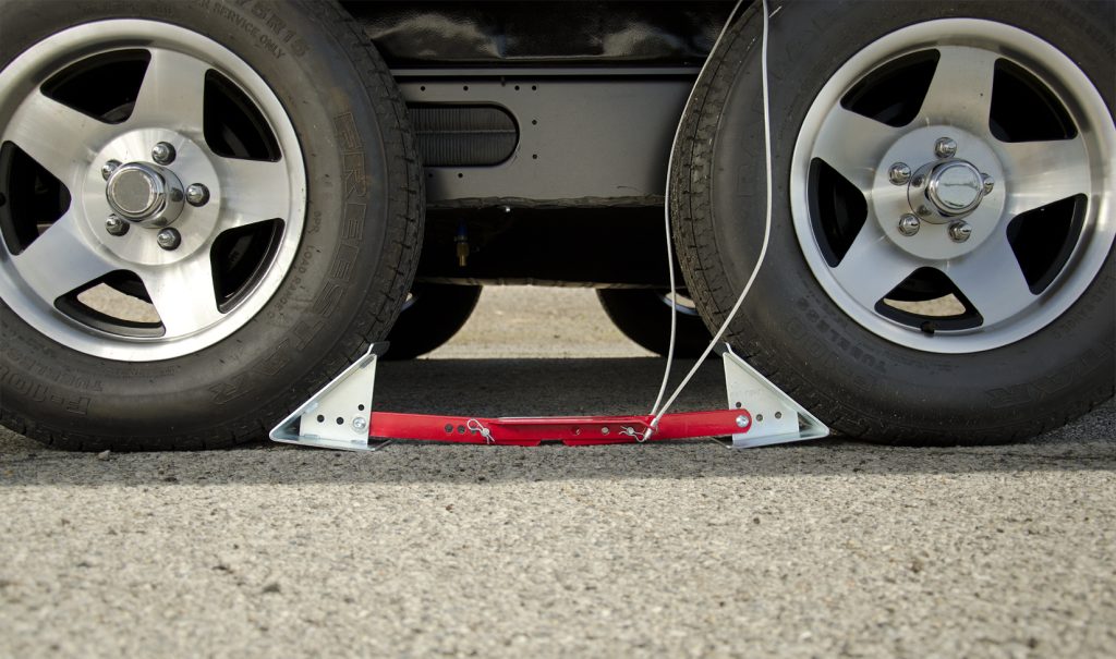 A single-axle utility trailer parked on a concrete driveway with two wedge wheel chocks placed tightly against the front and rear of the same tire