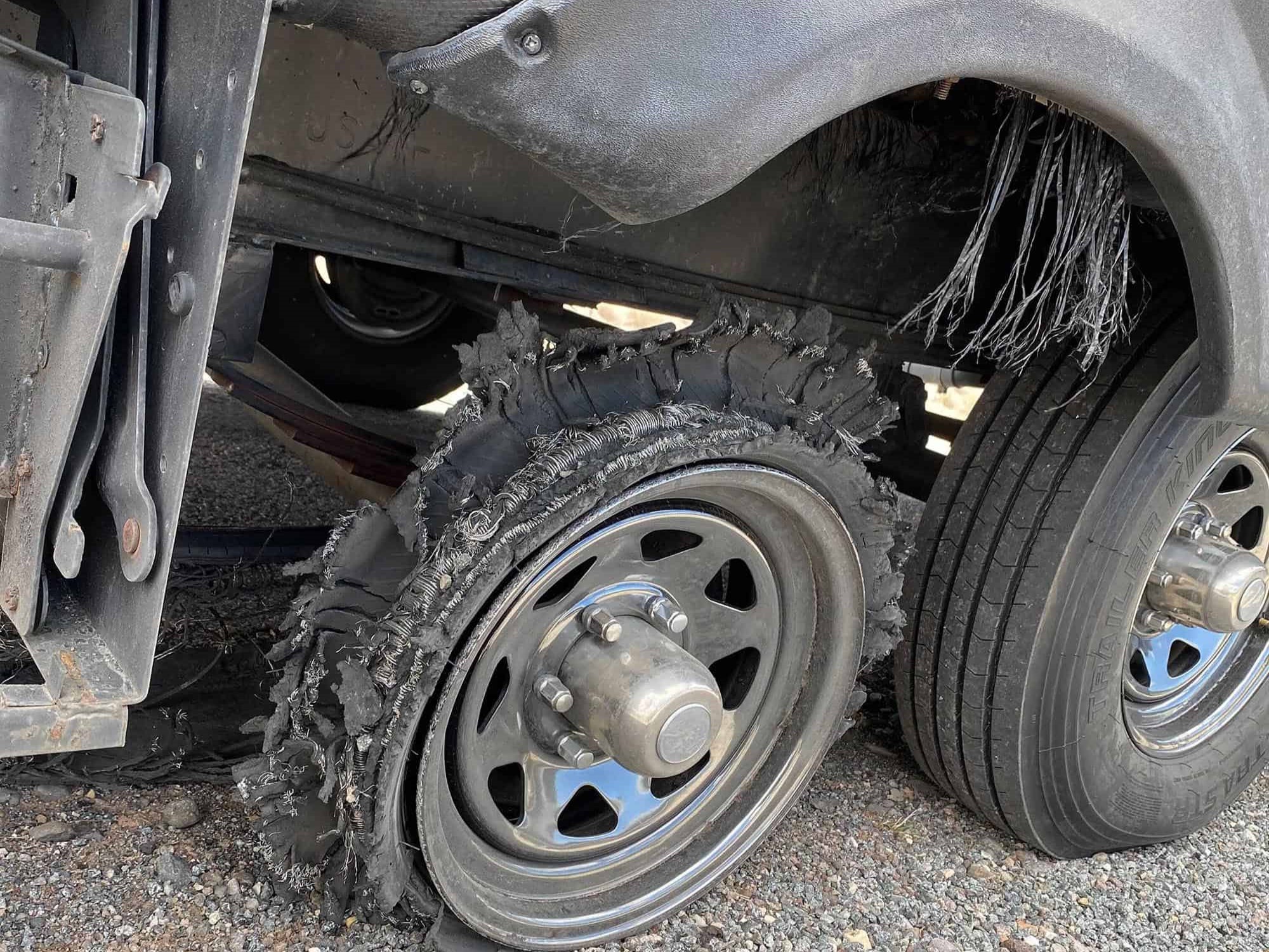 A travel trailer stopped on the right shoulder of a highway with a shredded trailer tire and rubber debris near the wheel well in daylight
